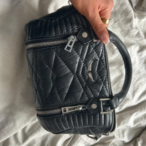 Svart handväska från Zadig & Voltaire - Säljer en svart quiltad handväska från Zadig & Voltaire med silverfärgade detaljer och dubbla dragkedjor. Väskan har ett stilrent mönster, handtag upptill och logga framtill. Perfekt storlek man får plats med allt från idrottskläder till smink trots att den är liten. Model: Sunny nano bag. Hör gärna av er vid frågor!! 😇 #zadig.  Band ingår!!