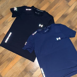 Under Armour t-shirt i svart och marinblå - Två snygga t-shirts från Under Armour, en i svart och en i marinblå. Båda har korta ärmar och logga på bröstet samt textdetaljer på ärm och nederkant. Perfekt för träning eller sportiga outfits.