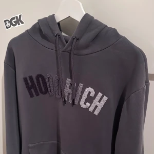 Grå hoodie från Hoodrich - Säljer en grå hoodie från Hoodrich med broderad logga i svart och vitt på bröstet. Tröjan har huva med dragsko och en klassisk känguruficka framtill. Perfekt för en avslappnad och street-inspirerad stil denna hoodie säljs ej längre 