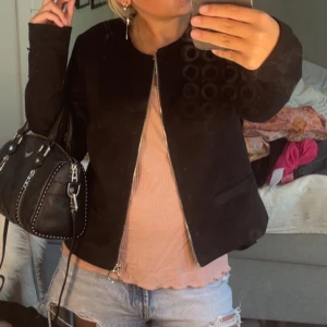 Svart manchesterjacka med dragkedja - Snygg svart manchesterjacka med rak passform och silverfärgad dragkedja framtill. Jackan har rund halsringning och diskreta fickor på sidorna. Perfekt för dig som gillar en stilren och enkel look.