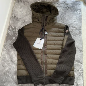 Olivgrön Moncler pufferjacka med huva - Snygg olivgrön pufferjacka från Moncler med stickade mörkgröna ärmar och detaljer. Jackan har huva, dragkedja framtill och Moncler-logga på ärmen. Perfekt för dig som vill ha en unik och stilren jacka med både quiltad och stickad känsla.