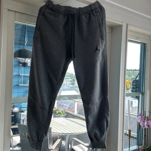 Mörkgrå Jordan mjukisbyxor - Snygga mörkgrå mjukisbyxor från Jordan med elastisk midja och snörning. Byxorna har ribbade muddar vid bensluten och två sidofickor. Ikonisk Jumpman-logga broderad på ena benet. Perfekta för chill eller träning.