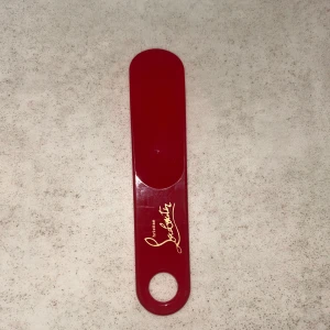 Röd skohorn från Christian Louboutin - Rött skohorn i plast från Christian Louboutin med ikonisk guldfärgad logga. Perfekt accessoar för att ta på sig dina favoritskor med stil. Smidig storlek och hål för enkel upphängning.