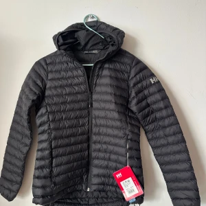 NY Svart lättviksjacka Helly Hansen XS - Women's Sirdal Hooded Insulated Jacket från Helly Hansen är en syntetfodrad jacka för dam. Jackan är fylld med återvunnen ECOLoft™ Air SR och har ett högt värme-vikt-förhållande. Den låga vikten och en avslappnad passform ger dig större förmåga att röra dig i naturen under skiftande förhållanden. Den smarta syntetiska isoleringen bildar till skillnad från traditionell dunfyllning inga klumpar när den är våt,. Yttermaterialet är ripstop som är utvecklat för att hålla. 