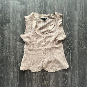 Beige blus med spets  - Supersöt beige blus med smala vertikala ränder, knappar framtill och spetsdetaljer längs krage, ärmhål och nederkant. Ärmlös modell med v-ringning och romantisk känsla. Perfekt mjuk beige färg snygg till Jeans eller linnebyxor 
