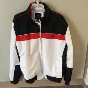 Vindjacka från U.S. Polo Assn, svart/vit/röd - Sportig vindjacka från U.S. Polo Assn. i svart, vitt och rött. Jackan har dragkedja, ståkrage med inbyggd luva. Broderad logga framtill samt 'USA' broderat på ryggen. Snygga kontrastpaneler och ribbade muddar ger en clean look. Perfekt för dig som gillar streetstyle.