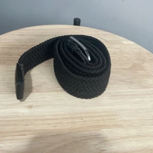 Svart flätat bälte med silverfärgat spänne - Snyggt svart flätat bälte med silverfärgat metallspänne och detaljer i skinn. Perfekt accessoar för att lyfta din outfit med en stilren och modern touch. Bältet har en flexibel design tack vare det flätade materialet.