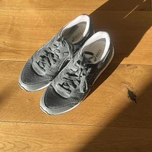 Grå sneakers från Asics - Snygga grå sneakers från Asics med vita detaljer och klassisk logga på sidan. Ovandelen är i mesh och syntet för bra andningsförmåga och komfort. Skorna har snörning och en rejäl, mönstrad sula som ger bra grepp. Perfekta för dig som gillar sportig stil.