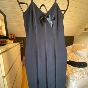 Mörkblå kort klänning med rosett - Snygg mörkblå kort klänning med smala axelband och v-ringning. Framtill finns en söt satinrosett och dekorativa vertikala band. Klänningen har en figurnära passform och dragkedja i ryggen. Perfekt för dig som gillar stilrena detaljer. inköpt på sellpy med prislapp kvar. Aldrig använd. 