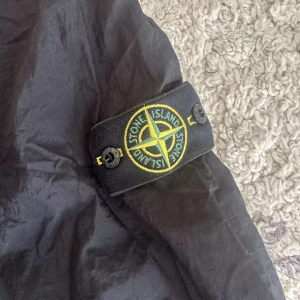 Svart overshirt från Stone Island - Svart overshirt från Stone Island med klassisk krage, dragkedja framtill och ikonisk patch på ärmen. Lätt och stilren jacka i syntetmaterial, perfekt för lager-på-lager. Passar dig som vill ha en clean och modern look.
