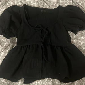 Svart blus med puffärm från Gina Tricot - Supersöt svart blus från Gina Tricot i storlek XS. Modellen har puffiga korta ärmar, knytning framtill och lätt rynkad struktur i tyget. Blusen har en flowy passform och är perfekt för dig som gillar trendiga detaljer och en feminin look.