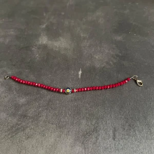 Rött pärlarmband med öga - Snyggt armband med små röda pärlor och ett evil eye-detalj i mitten. Armbandet har silvriga detaljer och ett smidigt spänne i metall. Perfekt accessoar för att ge din outfit lite edge och färg.