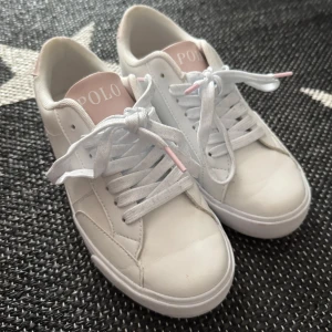 Vita sneakers från Polo Ralph Lauren - Snygga vita sneakers från Polo Ralph Lauren med ljusrosa detaljer på plös, häl och logga. Skorna har klassisk rund tå, platt sula och vita snören med rosa tippar. Ovandelen är i skinn och textil, perfekt för en clean och fräsch look. Använda ett fåtal gånger därav tecken på användning. 
