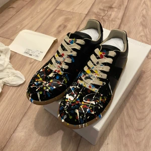Maison Margiela sneakers med färgstänk - Unika svarta sneakers från Maison Margiela med coola färgstänk i blått, rött, gult och vitt. Skorna har beige snörning, gummisula och är tillverkade i skinn. Perfekta för dig som vill sticka ut med en konstnärlig vibe.