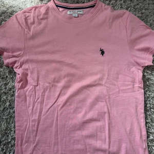 Rosa t-shirt från U.S. Polo Assn. - Snygg rosa t-shirt från U.S. Polo Assn. med klassisk rund hals och broderad logga på bröstet. Skön passform och tillverkad i mjuk bomull, perfekt för en avslappnad stil.