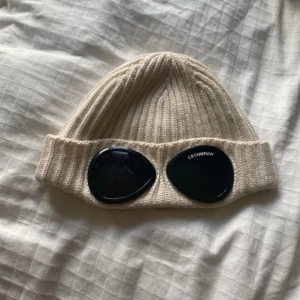 Beige C.P. Company goggle-mössa - Säljer en beige ribbstickad mössa från C.P. Company med de ikoniska svarta goggle-linserna framtill. Mössan är tillverkad i ett mjukt och värmande material och har en uppvikt kant. Perfekt för dig som vill sticka ut med streetwear-vibe. Har en liten pricka men syns inte när man har på sig den 