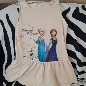 Frozen klänning (barn) - Supersöt beige ärmlös klänning med motiv av Elsa, Anna och Olaf från Frozen samt texten 'Family Forever'. Klänningen har volang nertill och är perfekt för alla som älskar Frost. Mjuk och skön bomullskänsla. Jätte fint skick, storlek 134/140 för 8-10 år