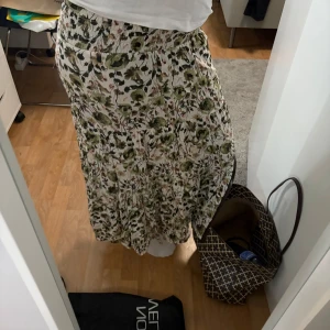 Blommig långkjol med volang - Supersöt långkjol med blommigt mönster i grönt, beige och svart på vit botten. Kjolen har bred resår i midjan och volang nertill för en flowy känsla. Perfekt för dig som gillar en avslappnad och romantisk stil. Kommer inte ihåg vart jag köpt den och har klippt bort lappen💕