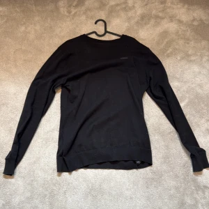 Svart långärmad tröja från Lindberg - En stilren svart långärmad tröja från Lexington med rund halsringning. Tröjan är i mjuk bomull och har en diskret logga på bröstet. Perfekt för dig som gillar en clean och enkel look. Passar till allt och är superbekväm.