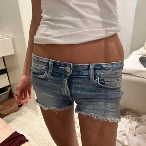 Lågmidjade jeansshorts  - Snygga lågmidjade jeansshorts i ljusblå denim med slitna, fransiga kanter och klassisk femficksmodell. På ena bakfickan finns en broderad fjäril som ger shortsen en unik touch. Perfekta för sommaren och riktigt trendiga.