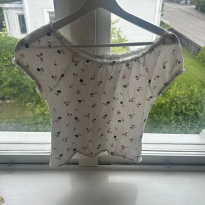 Vit blommig croppad topp - Supersöt vit croppad topp med små blommor i blått och rött. Toppen har korta ärmar, volangkant vid hals och nederkant samt en lätt rynkad struktur. Perfekt för en somrig look!