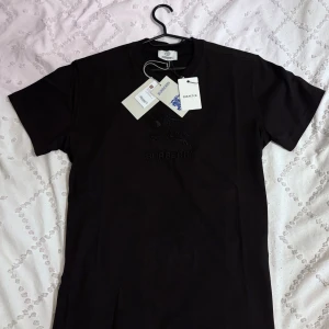 Burberry T-shirt  - Svart t-shirt från Burberry i 100% bomull med korta ärmar och rund hals. Framsidan har ett snyggt svart broderat Burberry-motiv och logga. Klassisk passform och exklusiv känsla, perfekt för dig som gillar stilrena designer.
