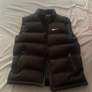 Svart dunväst  - Säljer en svart dunväst  med hög krage och dragkedja framtill. Västen har två fickor med dragkedja och en stilren, vadderad design med Nike-logga på bröstet. Perfekt för lager-på-lager och streetwear.