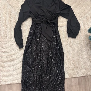 Svart långklänning med paljetter - Svart långklänning med glittrig paljettkjol och långärmad överdel i satinliknande material. Snygg knytning i midjan och v-ringad hals. Perfekt för dig som vill sticka ut på festen!