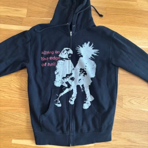 Svart hoodie Works of Madness M - Svart hoodie med dragkedja från Works of Madness. Framsidan har ett stort tryck i grått och vitt av två punkare samt texten 'sitting on the edge of hell' i rött. Huva med snörning och känguruficka. Tillverkad i USA, mjuk insida.