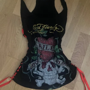 Ed Hardy svart linne med dödskalle - Svart linne från Ed Hardy med ikoniskt dödskalle- och hjärtmotiv framtill, dekorerat med glittrande stenar och röda samt gröna detaljer. Sidorna har röda snörningar som ger en edgy look. Tryck i guld och rött både fram och bak. Perfekt för dig som gillar statement-plagg.