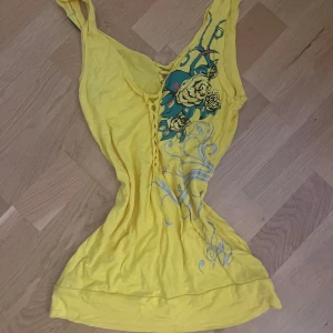 Gul linnetopp med tryck och snörning - Gul linnetopp med smala axelband och coolt tryck med blommor och mönster i grönt och grått. Toppen har snörningsdetalj framtill och glittriga stenar på ryggen. Perfekt för dig som vill sticka ut med färg och detaljer.