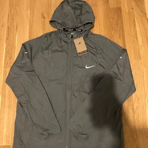 Grå nike Miller jacka - Snygg grå vindjacka från Nike med huva och dragkedja framtill. Jackan har en stilren look med Nike-logga på bröstet och elastiska muddar vid ärmsluten. Perfekt för blåsiga dagar och enkel att matcha med det mesta.