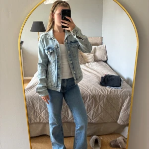 Ljusblå jeansjacka med klassisk passform - Säljer en ljusblå jeansjacka med klassisk krage, knappar framtill och två bröstfickor med lock. Jackan har långa ärmar och raka sömmar, perfekt för en chill och stilren look. Passar till allt och är ett måste i garderoben.