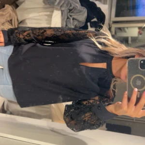 Svart blus med spetsärmar och slits - Säljer en svart blus med transparenta spetsärmar och snygg slits vid halsen. Ärmarna har blommigt spetsmönster och knäppning vid mudden. Perfekt för dig som vill ha en stilren men ändå unik topp.