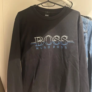 Svart tröja från Hugo Boss - Svart tröja från Hugo Boss med stor logga i vitt och blått framtill. Denna tröja är ganska sällsynt och fantastiskt fin, den är också i mycket bra skick