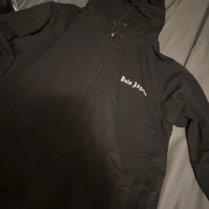Svart vindjacka från Palm Angels - Svart vindjacka från Palm Angels med huva och dragkedja framtill. Jackan har logga broderad på bröstet och praktiska fickor. Perfekt för dig som gillar streetwear och vill ha en clean look med snygga detaljer. Storlek L/XL