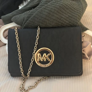 Svart axelväska Michael Kors - Snygg svart axelväska från Michael Kors med guldfärgad kedja och stort MK-emblem framtill. Väskan har diskret MK-mönster i materialet och invändigt foder med logga. Perfekt storlek för det viktigaste och ger en lyxig känsla till din outfit. Den är äkta!