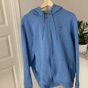 Blå hoodie med dragkedja från Lacoste - Säljer en blå hoodie från Lacoste med dragkedja framtill och klassisk krokodillogga på bröstet. Tröjan har huva med snörning, fickor på sidorna och är tillverkad i mjuk bomullsblandning. Perfekt för chill dagar eller när du vill ha en clean streetstyle. Byxor tillhör!