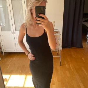 Svart långklänning med smala axelband - En stilren svart långklänning med smala axelband och figurnära passform. Klänningen har en enkel och tidlös design som ger en snygg siluett. Perfekt för dig som gillar minimalistisk stil och vill ha något som känns både bekvämt och trendigt.