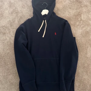 Mörkblå hoodie från Polo Ralph Lauren - Säljer en mörkblå hoodie från Polo Ralph Lauren med klassisk röd logga broderad på bröstet. Tröjan har huva med vita snören och en stor magficka. Perfekt för dig som gillar stilren och sportig look.