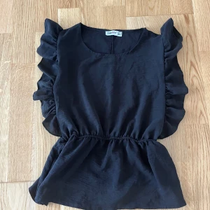 Svart blus med volangärmar från Sheryy - Supersöt svart blus från Sheryy med volangdetaljer längs ärmarna och markerad midja.  Har använts bara ett par gånger 