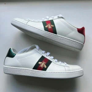Gucci Ace vita sneakers - Gucci Ace sneakers i vitt skinn med klassiska röd-gröna ränder på sidorna och broderad guldfärgad bi. Skorna är helt nya. Storlek 40,5 men passar 1 storlek större.