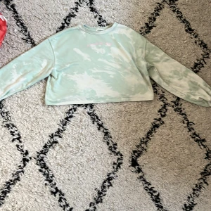 Mintgrön croppad sweatshirt - Perfekt för yngre barn! Säljer en croppad sweatshirt i mintgrönt och vitt tie dye-mönster. Tröjan har rund halsringning och långa ärmar med avslappnad passform. Perfekt för dig som gillar en chill och trendig stil.