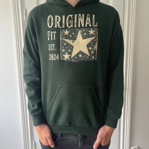 Mörkgrön hoodie ifrån Scndi med stjärntryck - Snygg mörkgrön hoodie med stor beige stjärna och texten 'ORIGINAL FIT EST. 2024' framtill. Klassisk känguruficka och huva. Perfekt för dig som gillar streetstyle och vill ha en chill look.
