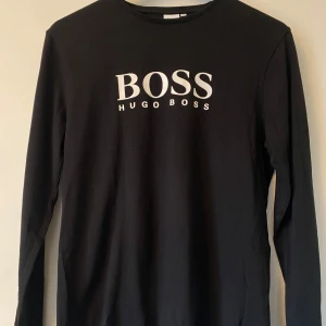 Svart långärmad tröja Hugo Boss - Svart långärmad tröja från Hugo Boss med stor vit logga på bröstet. Tröjan är i mjuk bomull med en klassisk rund halsringning och har en clean, stilren look som passar perfekt till jeans eller chinos.