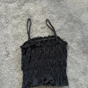 Svart volangig linnetopp - Supersöt svart linnetopp med smala axelband och volangdetaljer både upptill och nedtill. Toppen har en smockad struktur som ger en snygg och bekväm passform. Perfekt att styla med jeans eller kjol för en trendig look.