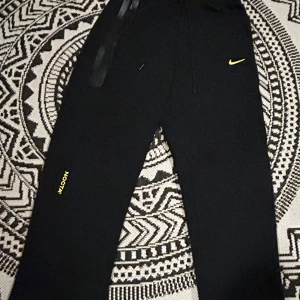 Svarta NOCTA x Nike mjukisbyxor - Svarta mjukisbyxor från Nike NOCTA med gul logga på benet och en avslappnad passform. Byxorna är tillverkade i mjukt material för maximal komfort. Perfekta för en avslappnad stil eller streetwear. Tveka inte på att höra av er vid minsta fundering.