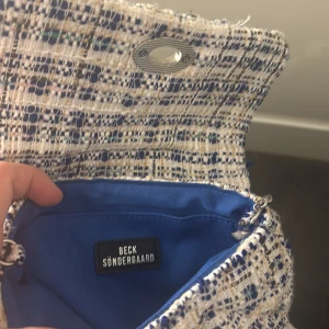 Rutig axelväska från Beck Söndergaard - Säljer en snygg axelväska från Beck Söndergaard i rutigt tyg med blå, beige och vita toner. Väskan har silverfärgad kedjerem och ett klassiskt vridlås framtill. Insidan är fodrad i blått tyg och materialet är mjukt och textiligt.