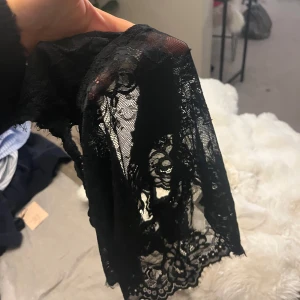 Svart spetsig linnetopp - Säljer en svart linnetopp i genombruten spets med blommigt mönster. Toppen har breda axelband och v-ringning, samt fina vågiga kanter nertill och vid axelbanden. Perfekt för dig som gillar en feminin och trendig look💕💕