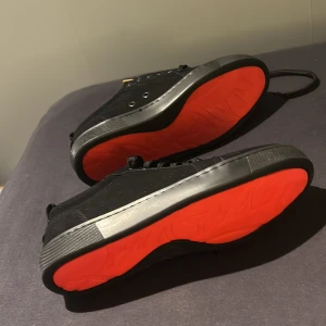 Svarta Louboutin sneakers med röd sula - Snygga svarta sneakers från Christian Louboutin med ikonisk röd sula. Skorna har ovandel i mocka, svarta snören och diskret logga på sulan. Sulan är platt. Perfekta för dig som vill sticka ut med exklusiv streetstyle. I storlek 43